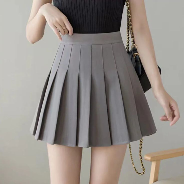 a woman is wearing a grey pleated mini skirt, black top, and a chain bag. she is standing with her right hand on her hip and her left hand is reaching out to the side. she is wearing a gold ring on her left hand. the photo is taken from the waist up. the woman's legs are visible.