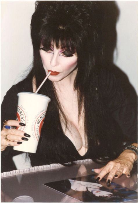 a photo of a dark haired woman with dark lipstick posing in a dark, gothic style. she is sitting at a table in a studio, with a cup in hand. she is dressed in all black, and her makeup is dramatic. she has long dark hair, and her eyes are lined with eyeliner. she is looking into the camera with a slightly mischievous smile.