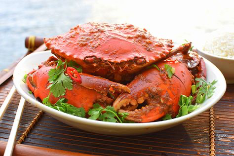 a closeup shot of a plate of chili crab, a popular singaporean dish. the crab is bright red from the chili sauce and is garnished with coriander. chopsticks and a bowl of rice are visible in the background. the setting appears to be outdoors on a bamboo placemat. the image emphasizes the vibrant color and texture of the food.