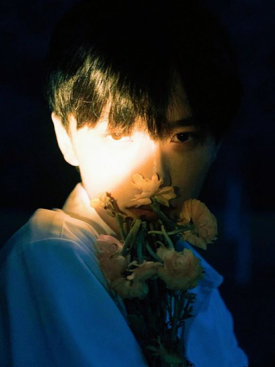 a young man with short, dark hair is partially illuminated by a dramatic spotlight against a dark background. he holds a bouquet of pale pink and yellow flowers in front of his face, obscuring part of his features. the lighting emphasizes the contrast between light and shadow, creating a moody and evocative atmosphere.