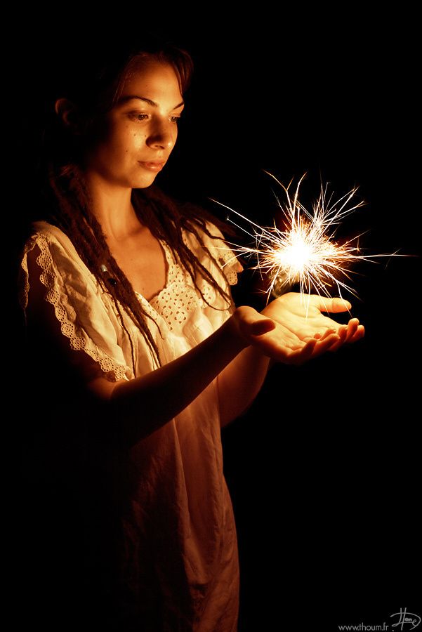 a young woman with dreadlocks is holding a sparkler in her hands while looking at it. she is wearing a white dress and has a serious expression on her face. the background is dark and the photo is lit by the sparkler.