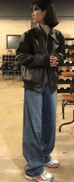 a woman in a black leather jacket and blue jeans stands in a room with wooden crates.  she is wearing white sneakers.  she is looking off to the side and has a casual, cool look.