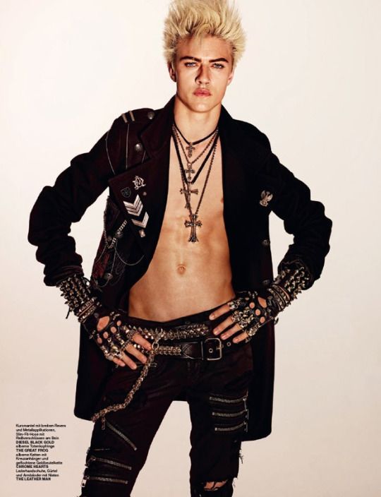 a male fashion model with blond, spiky hair is posing. he is wearing an open black jacket, exposing his torso, with several necklaces featuring cross pendants. he is also wearing black pants with zipper details, a studded belt with chains, studded gloves and wristbands, and multiple rings. he has his hands on his waist. the background is plain white.