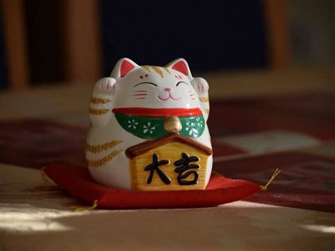 a closeup shot of a manekineko, a japanese beckoning cat figurine, sitting on a red cushion. the cat is predominantly white with gold and brown markings, wearing a green and red collar.  a small yellow houseshaped sign with japanese characters is attached to its chest. the background is blurred but shows a dark woodlike surface and a burgundycolored tabletop. the overall lighting and composition create a warm, inviting, and slightly mystical atmosphere.