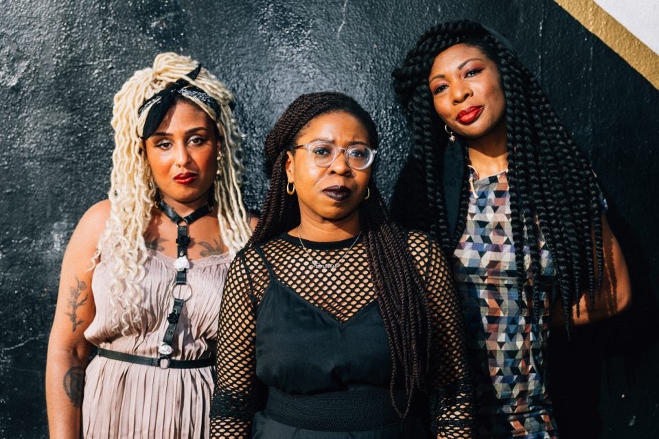 three women of color stand in front of a dark wall, posing for the camera. two of the women are wearing braids, one blonde and the other black. the third woman has her black hair in braids that are pulled back and away from her face. the woman on the left is wearing a tan and pink dress with black straps, and a black choker with white flowers and black trim. the woman in the middle is wearing a black shirt with a black fishnet top layered over it. the woman on the right is wearing a brightly colored patterned shirt. all of them are smiling, and they appear to be having fun.