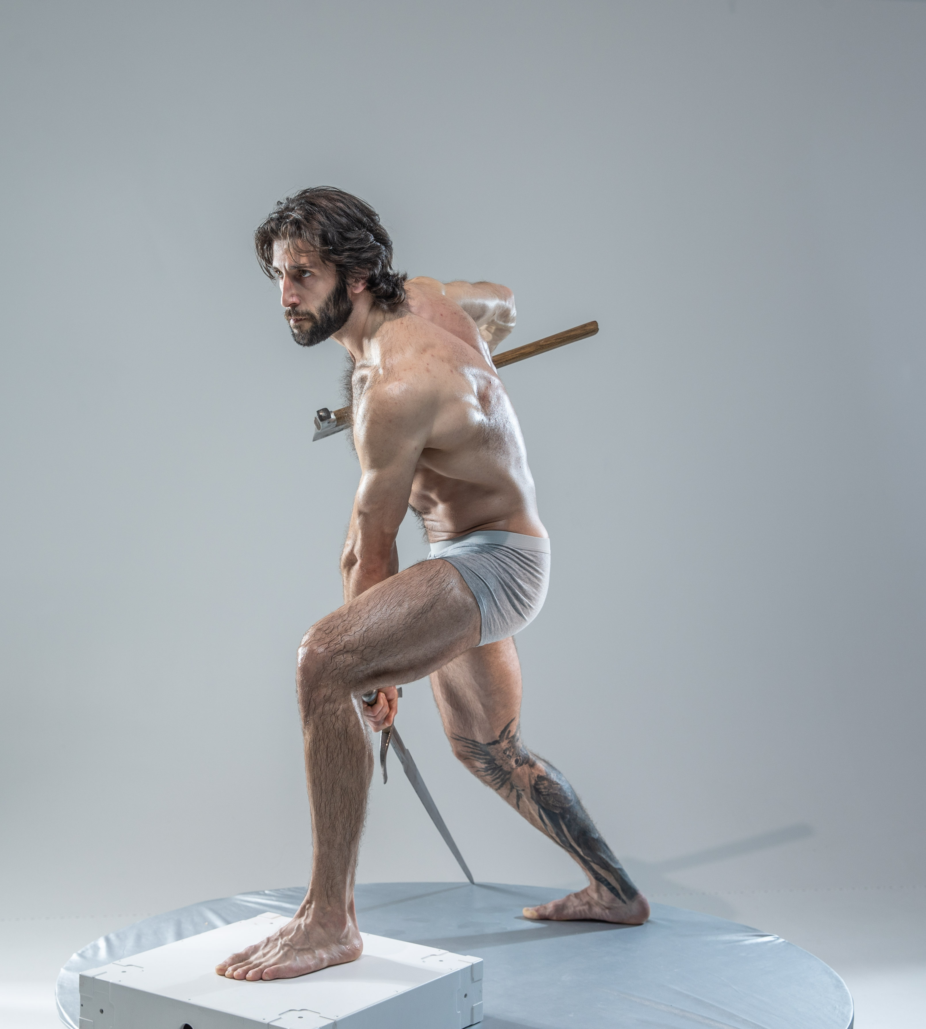 a muscular man with long hair and a beard stands in a dynamic warrior pose on a small white platform against a solid gray background. he is wearing only underwear and is holding a short sword in each hand. he is illuminated by soft studio lighting.