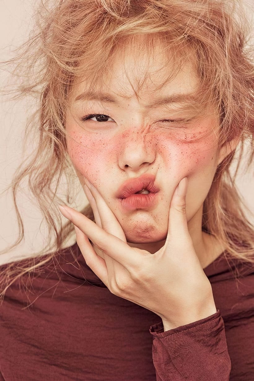 a closeup portrait of a young woman with messy, light blonde hair. she is making a playful, slightly silly facial expression, with rosy cheeks and visible freckles. her hands are gently cupping her face. she's wearing a maroon, longsleeved turtleneck top. the background is a simple, light beige, drawing focus to the subject's expressive features and unique style. the overall mood is lighthearted and fashionable.