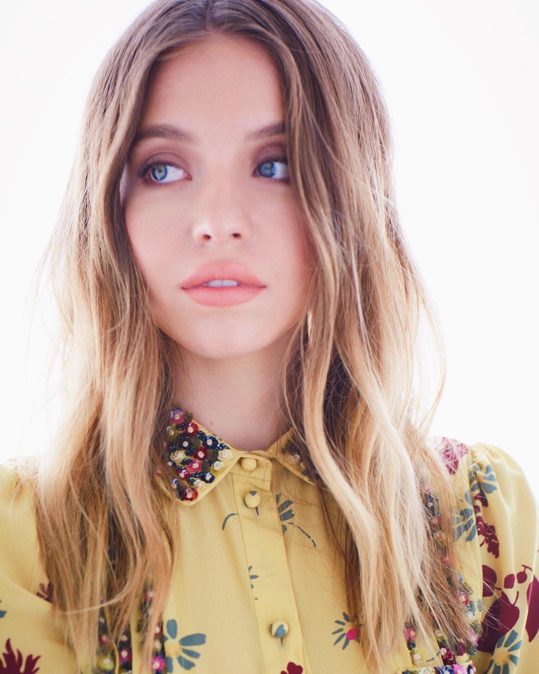 a closeup portrait of a young woman with long, wavy blonde hair. she is wearing a pastel yellow floral shirt with a beaded collar. the background is bright white. the overall mood is soft and elegant. the woman looks directly to her right.  her expression is serene. the focus is on the woman's face and upper body.