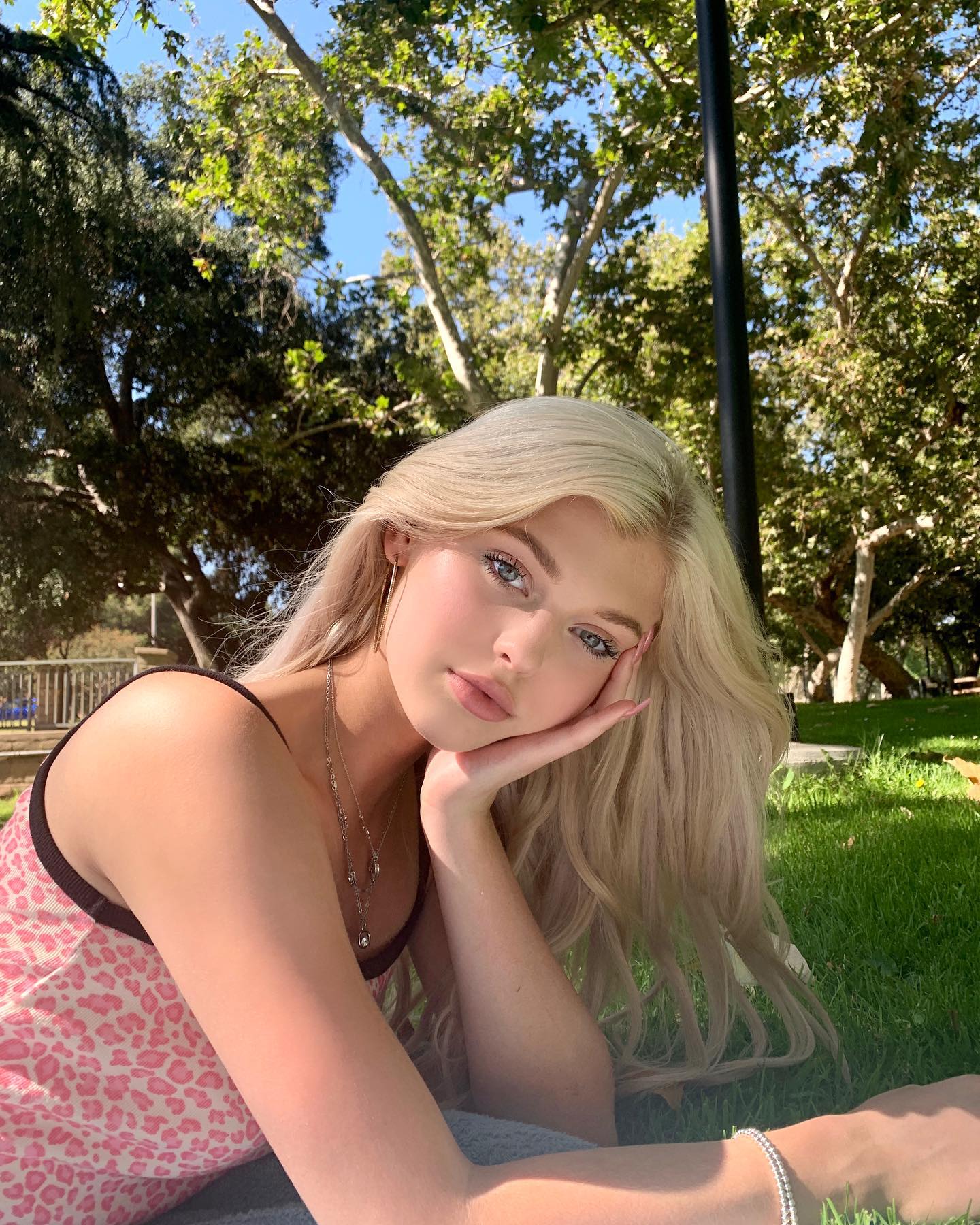 a woman with blonde hair and blue eyes is lying down in a park on the grass. she is wearing a pink leopard print top, earrings, a necklace, and a bracelet. she has her hand on her face and is looking at the camera. there are trees in the background. it is daytime.
