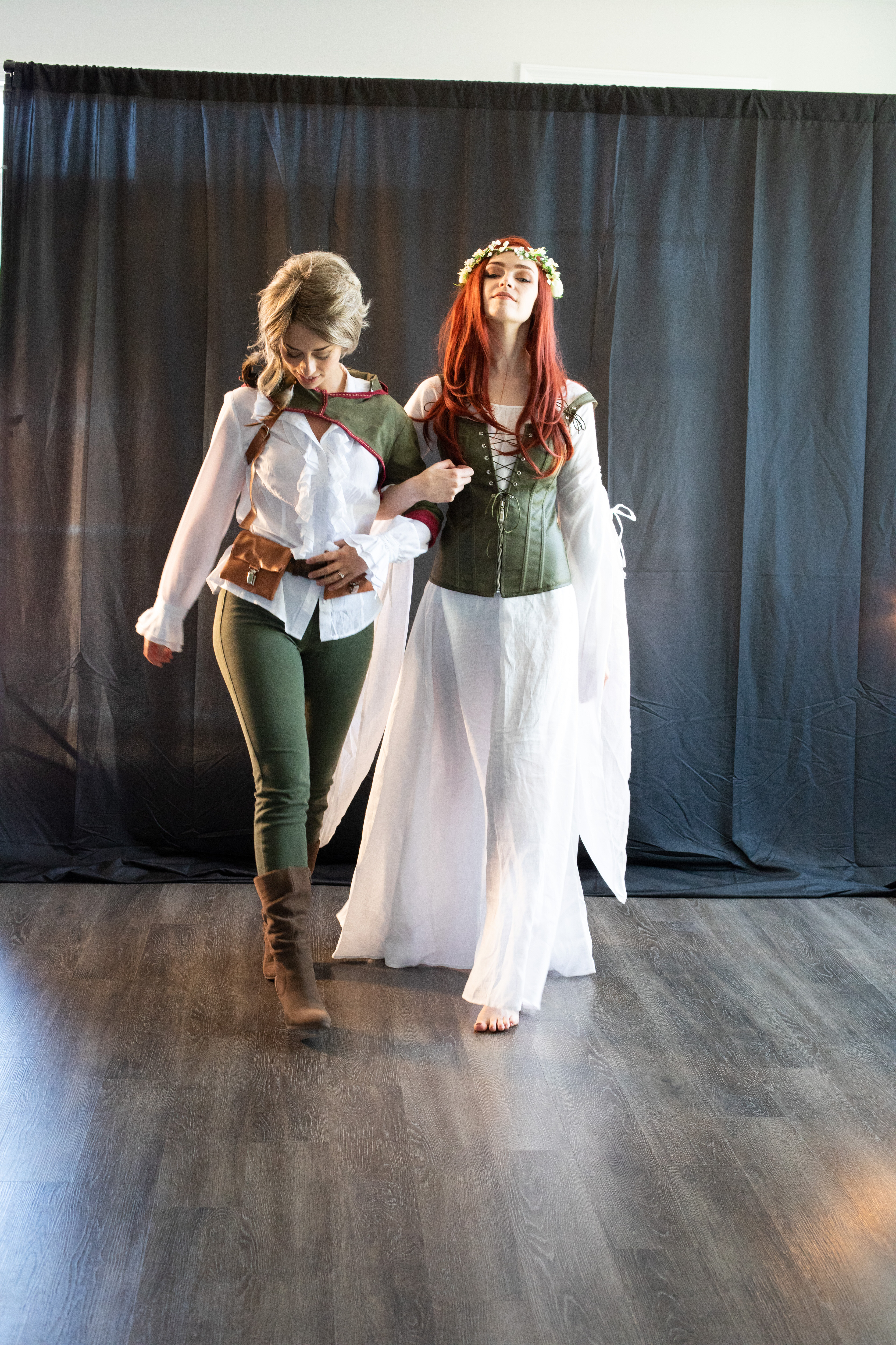 two women dressed in fantasy cosplay walk across a wooden floor against a black curtain backdrop. the woman on the left has blonde hair and is wearing a white top, green pants, and brown boots and belt. she is also wearing a brown wrist cuff on her right wrist. the woman on the right has red hair with a crown of flowers and is wearing a flowing white dress with a green corset.