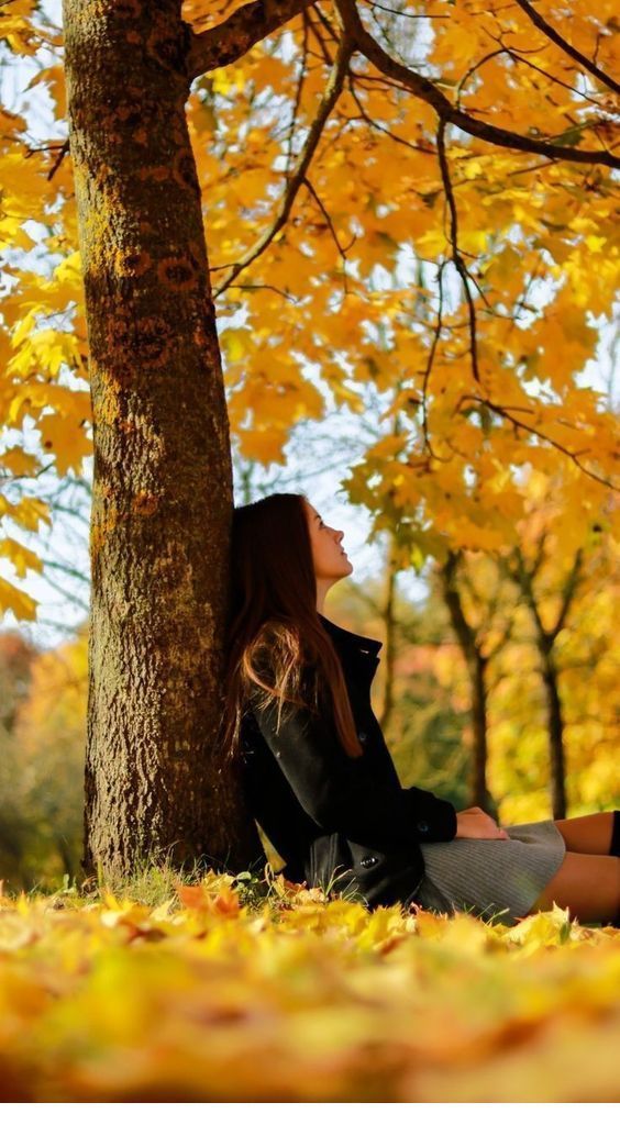 young woman with long brown hair is sitting on the ground, leaning against a large tree trunk. the tree is surrounded by vibrant yellow and orange autumn leaves. she is wearing a dark coat and a grey skirt. the scene is peaceful and serene, capturing the beauty of autumn. the sunlight filters through the leaves, creating a warm and inviting atmosphere. the image conveys a sense of calm and contemplation.