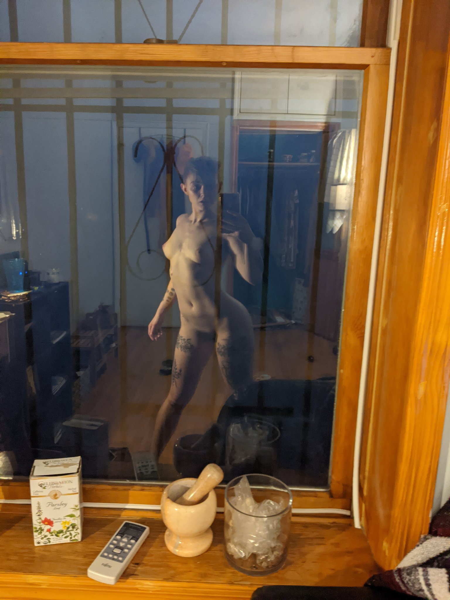 the image shows a reflection of a nude woman in a window mirror. the woman is standing and posing, her body is visible, with tattoos on her legs. the reflection is slightly distorted by the glass. the window has a wooden frame. on the windowsill are a floral tea box, a mortar and pestle, a glass with ice, and a remote control. a corner of a blanket is visible in the bottom right. the background shows a glimpse of a room's interior.