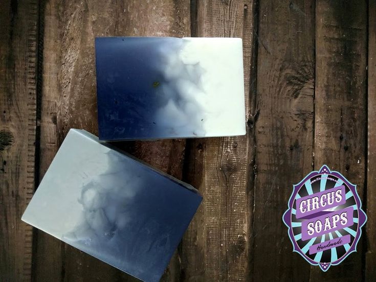 a closeup shot of two handmade soap bars on a wooden background. the soap bars are a gradient of white and blue, with a swirl pattern. there is a purple and turquoise label that says "circus soaps" in black letters. the label is also on a wooden background.