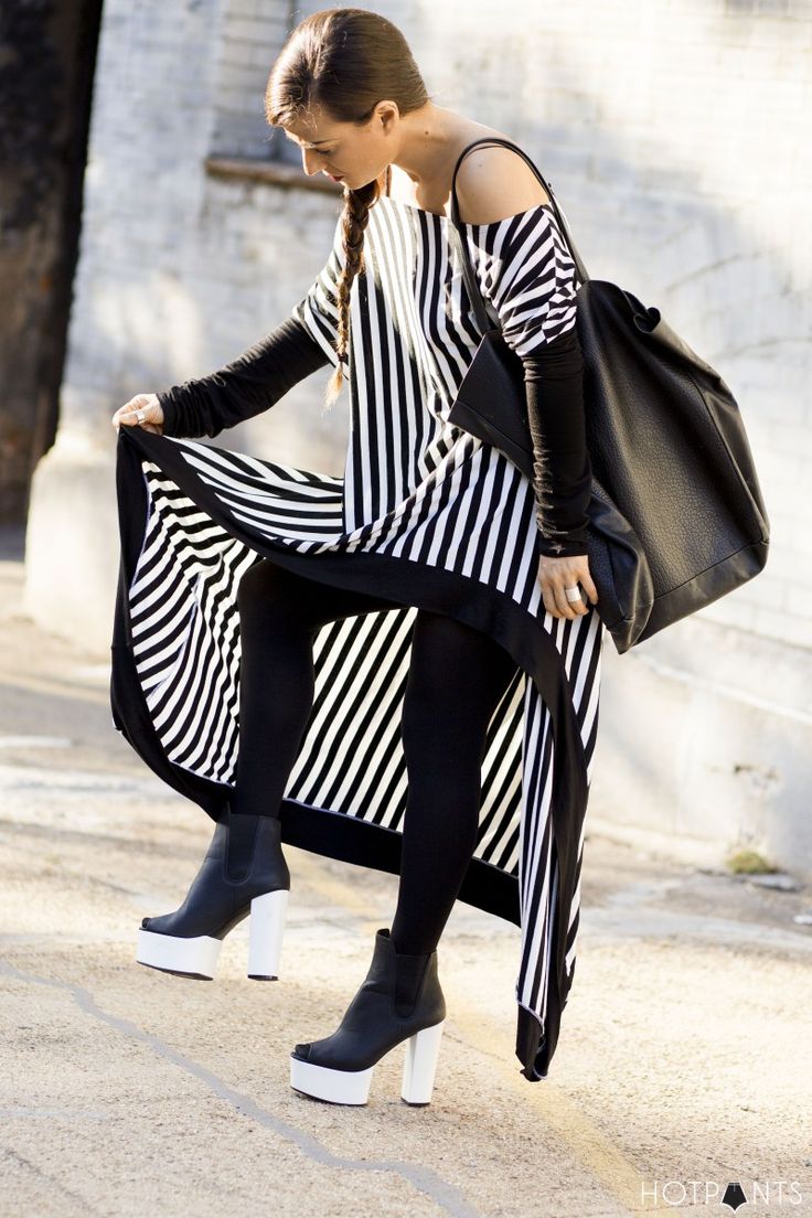 a woman is standing outside, holding up the edge of her black and white striped dress to reveal black leggings and platform boots. she has a black bag slung over her shoulder and her hair is in a braid. the dress has a oneshoulder design with a black longsleeved shirt underneath.