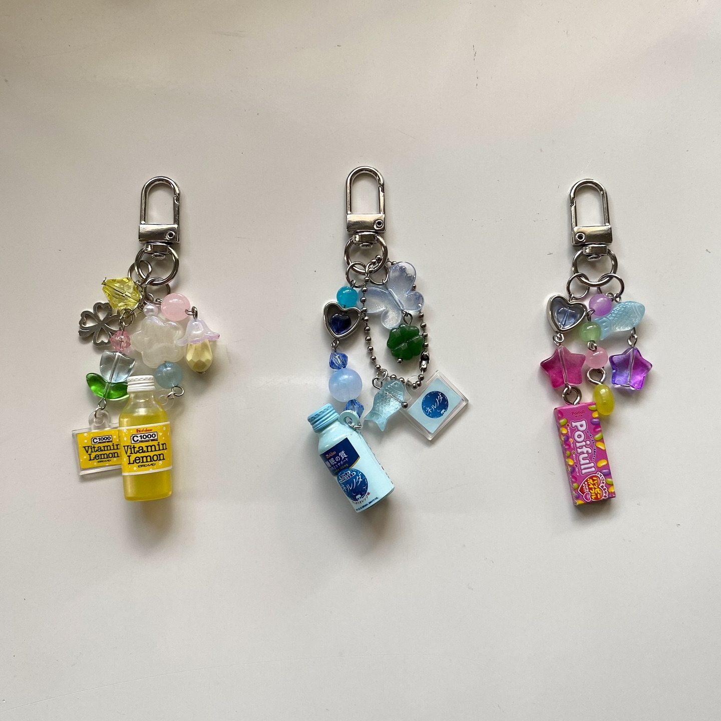 three keychains are arranged horizontally on a white surface. each keychain consists of a silver clasp and a variety of colorful charms, beads, and miniature items. the left keychain features a yellow vitamin lemon drink miniature with accompanying yellow and green charms. the middle keychain has a light blue bottled drink miniature and blue beads. the right keychain has a polfull candy miniature and pink and purple star and heart charms. all keychains use pastel colors and shapes like clovers, hearts, stars, and butterflies.