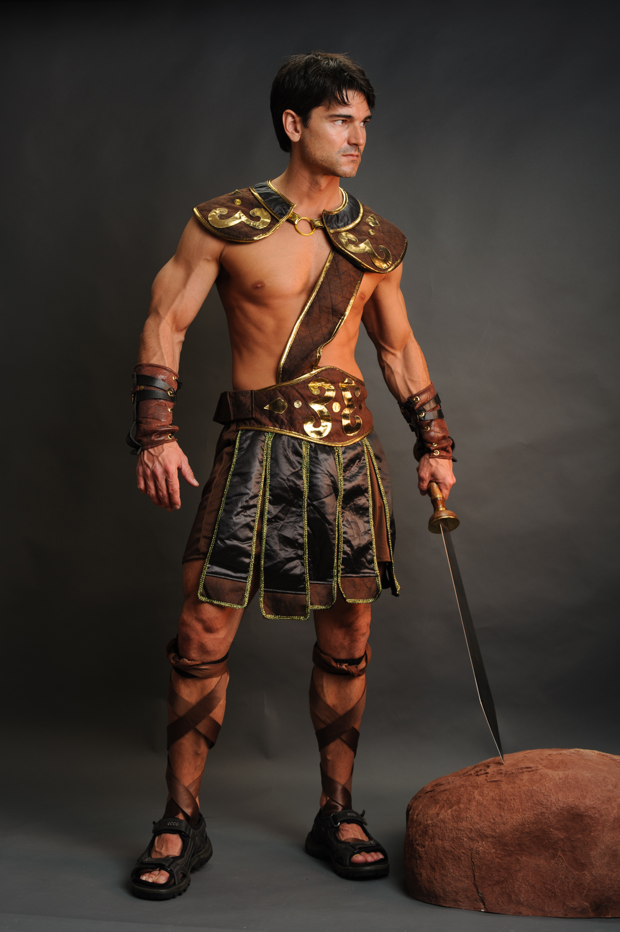 a muscular man dressed as a gladiator or warrior stands in front of a gray background with a sword in his hand. he is wearing armor, a short skirtlike garment, sandals, and arm wrappings. a brown rock or mound is near his feet.