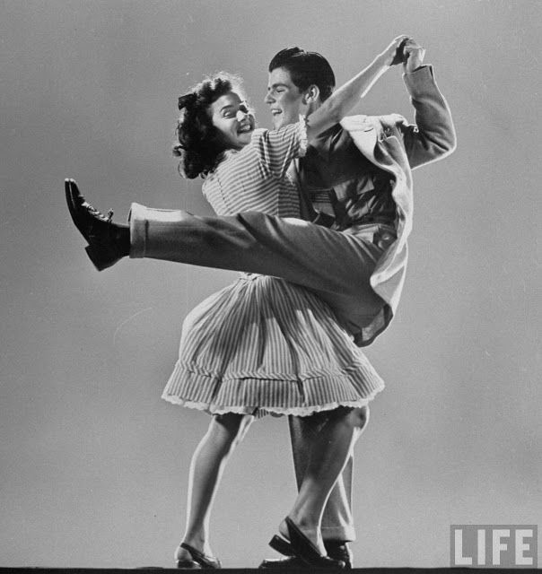 a young couple in 1950s attire dances joyfully, the man holding the woman's legs as they both smile and look at each other. the man is in a jacket and slacks, and the woman in a dress with a skirt. the photo is taken in a studio and is in black and white.