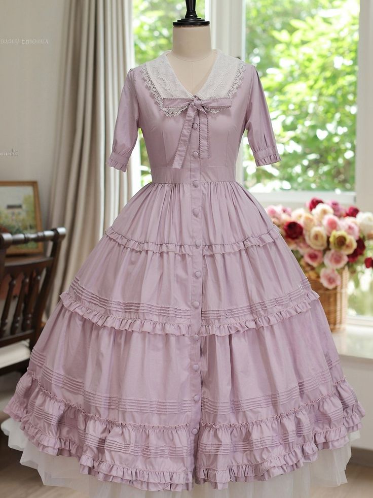a lavender vintagestyle dress is displayed on a mannequin. the dress has a full, tiered skirt with ruffles and a buttonfront bodice.  it features a white lace collar and short puffed sleeves, tied with a bow at the neckline. the dress sits on a  white petticoat, adding volume. the background shows a room with a window and a chair, with a bouquet of flowers to the right of the mannequin.