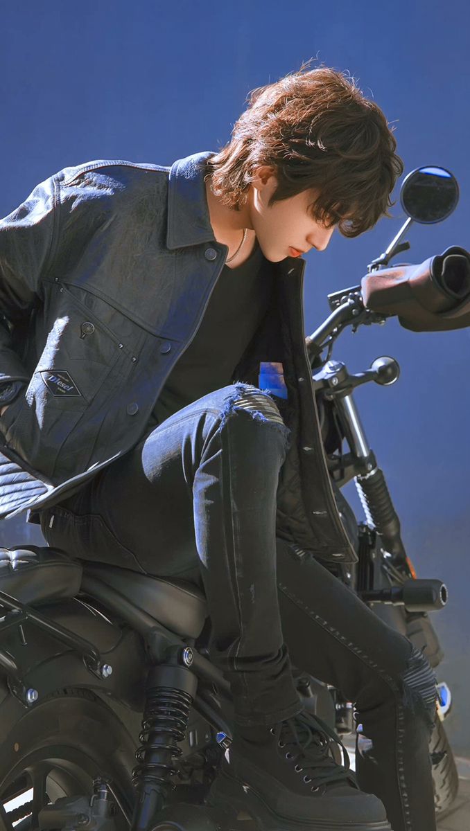 a young man with a trendy, shaggy haircut sits on a black motorcycle. he's wearing a denim jacket, black jeans with ripped knees, and black boots. the man is turned to the side, looking down with a contemplative expression. the background is a bright, clear blue sky, suggesting it's a sunny day. the overall image conveys a sense of coolness and effortless style.