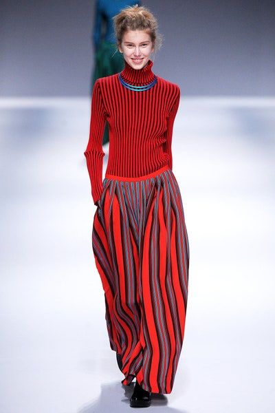 a model walks down a runway at a fashion show. she is wearing a red and black striped turtleneck sweater, a highwaisted skirt with red and black stripes, and a blue necklace. the skirt is voluminous and pleated. she has a bun hairstyle and is smiling. the background is a neutral color.