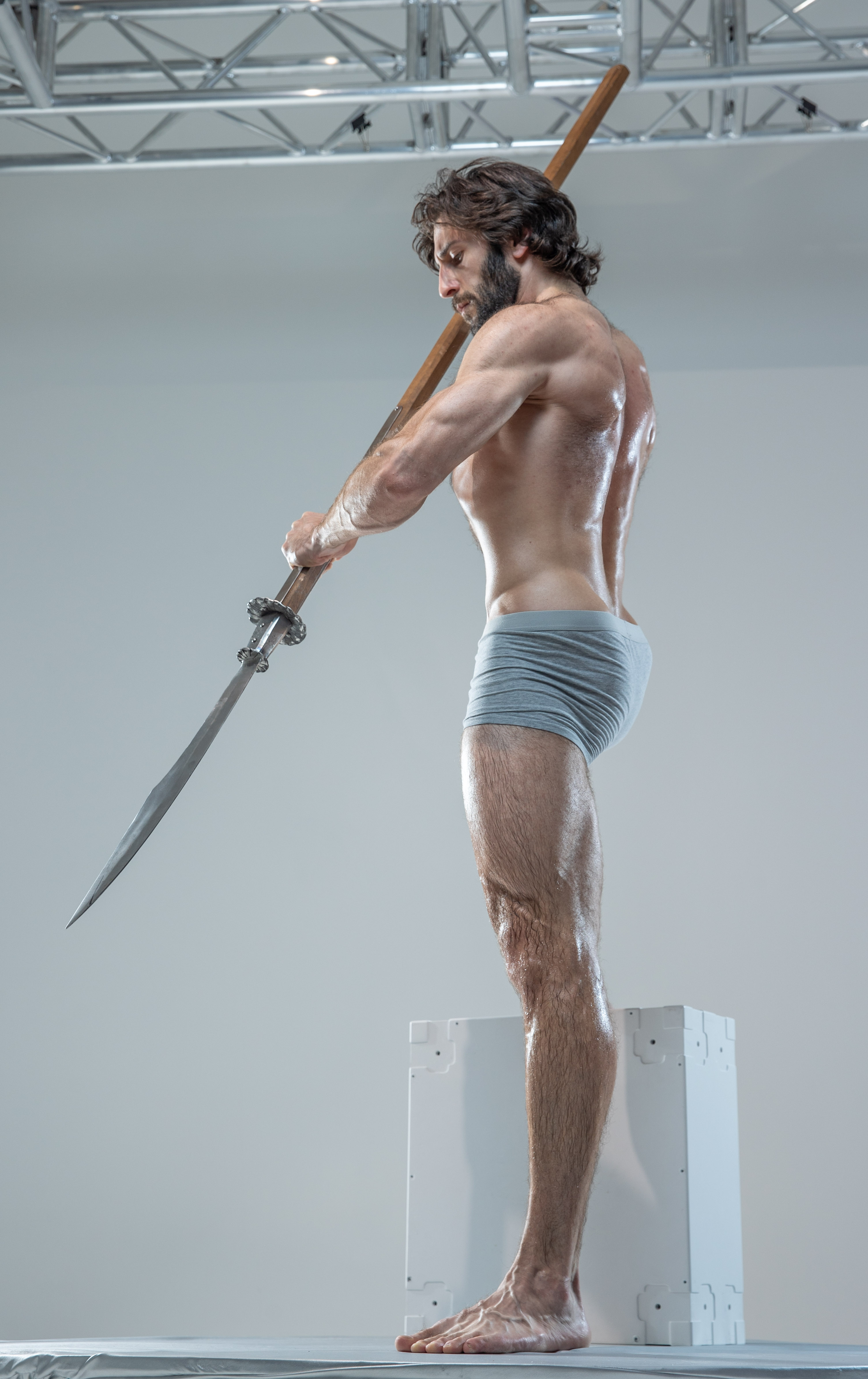 a muscular man with long hair and beard, wearing only gray underwear, stands in a contrapposto pose holding a spear.  he is illuminated with soft studio lighting against a white background. his pose and expression suggest a sense of strength, power, and determination.