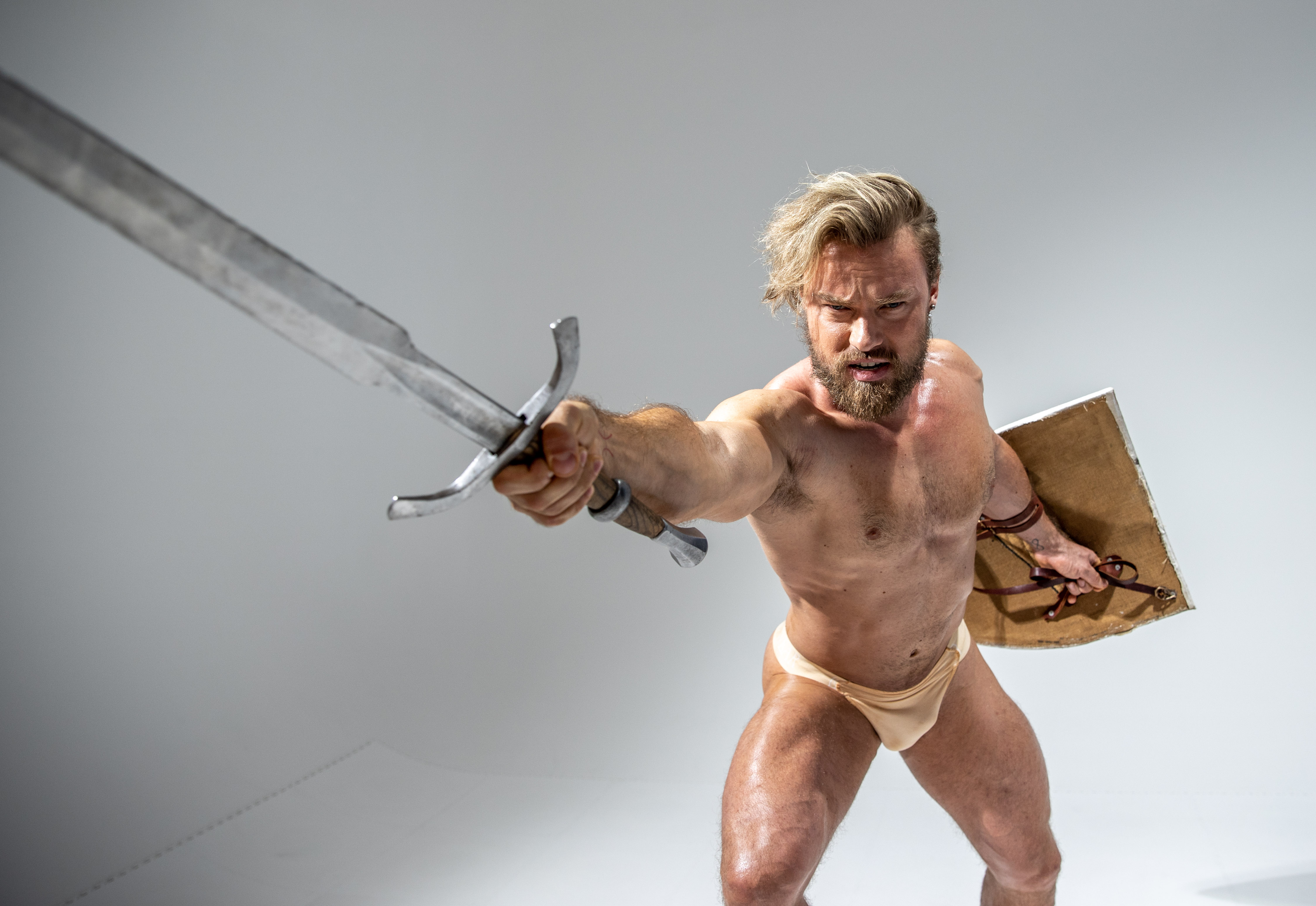 a muscular man with blonde hair and beard, dressed in beige underwear, is captured in a fighting stance against a plain white background. he wields a long, silver sword in his right hand, thrusting it forward towards the viewer. in his left hand, he holds a round, brown shield. the image is shot from a low angle, emphasizing the man's strength and power.