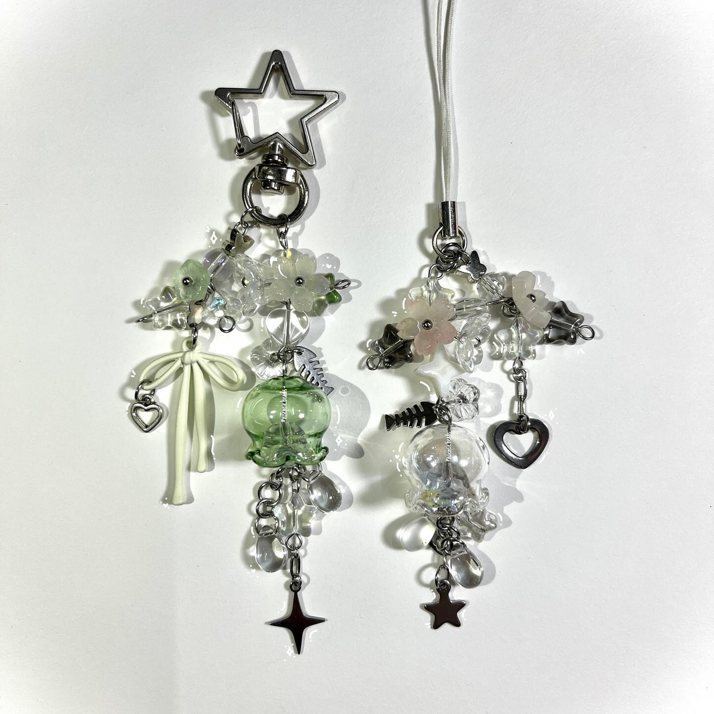 the image shows two handmade keychains or phone charms. one has a silver star clip, a white bow with a heart, a green glass bead, and a black star charm. the other has a white strap, a fishbone charm, and a silver star charm. both have flowers and clear beads.
