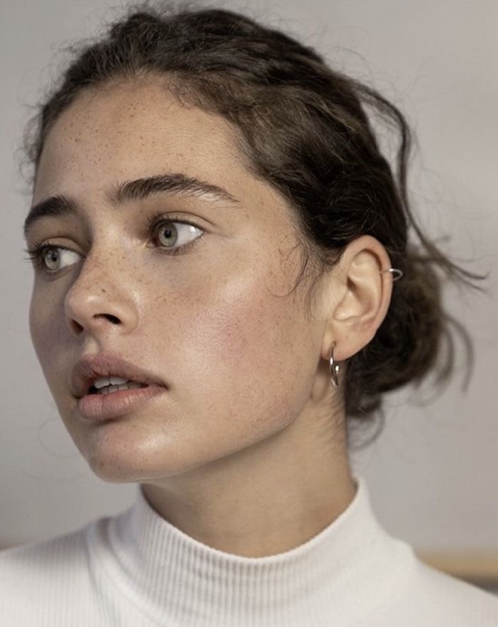 closeup portrait of a young woman with light skin and freckles. she has brown hair styled in a messy bun and is wearing simple silver hoop earrings. the woman is wearing a white turtleneck and has minimal makeup, showcasing her natural beauty. the background is a simple, neutral color. the focus is on her face, emphasizing her complexion and features.