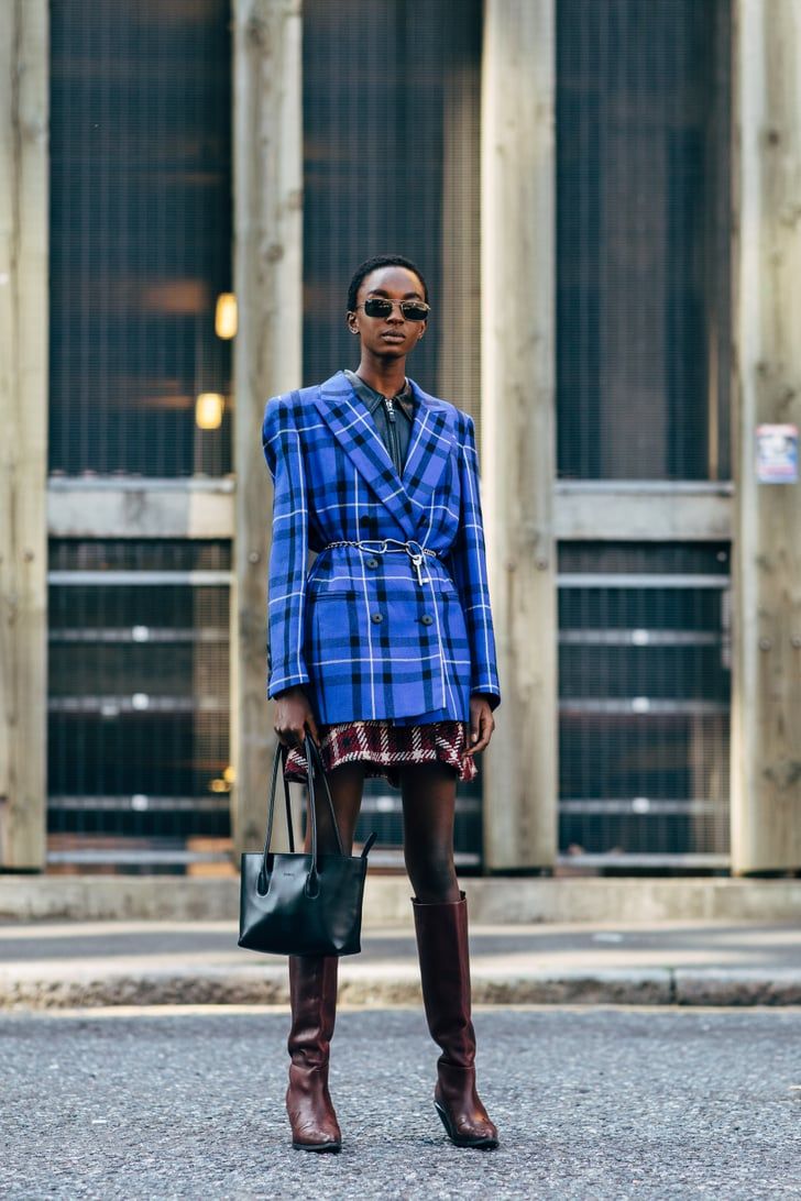 the image showcases a stylish woman, possibly a model, on a city street. she's wearing a striking blue and white oversized plaid blazer over a plaid mini skirt. the skirt is a slightly darker shade of red and burgundy.  she's accessorized with burgundy kneehigh boots, a black tote bag, and sunglasses. the background features a modern urban building with a gridlike facade. the overall style is chic, modern, and appropriate for autumn.