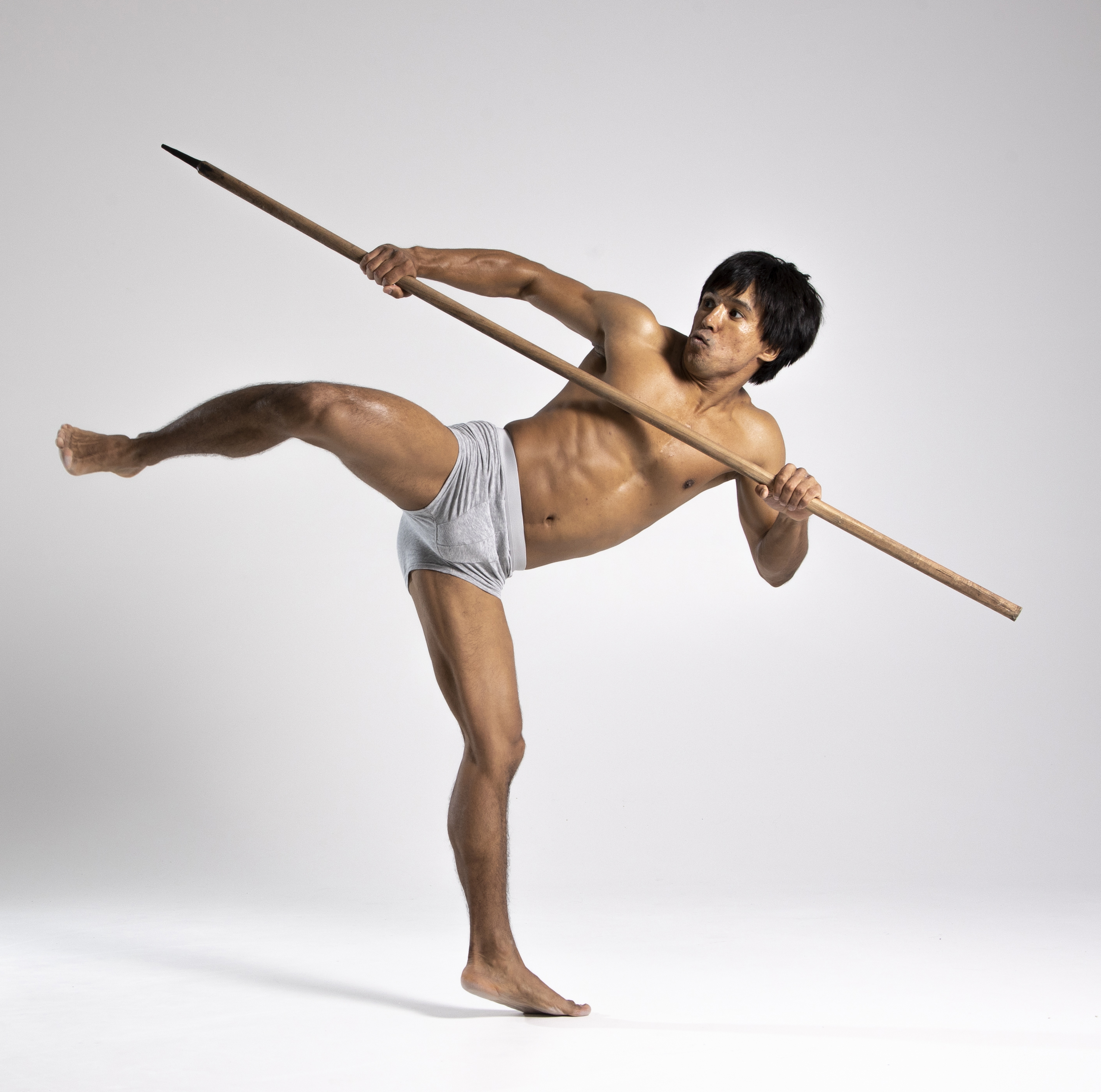 a muscular, shirtless man in gray underwear is holding a spear while balancing on one leg. he is in a martial arts pose, with one leg extended in front of him and the spear held horizontally across his body. he is barefoot and appears to be asian. the background is a plain, light gray.