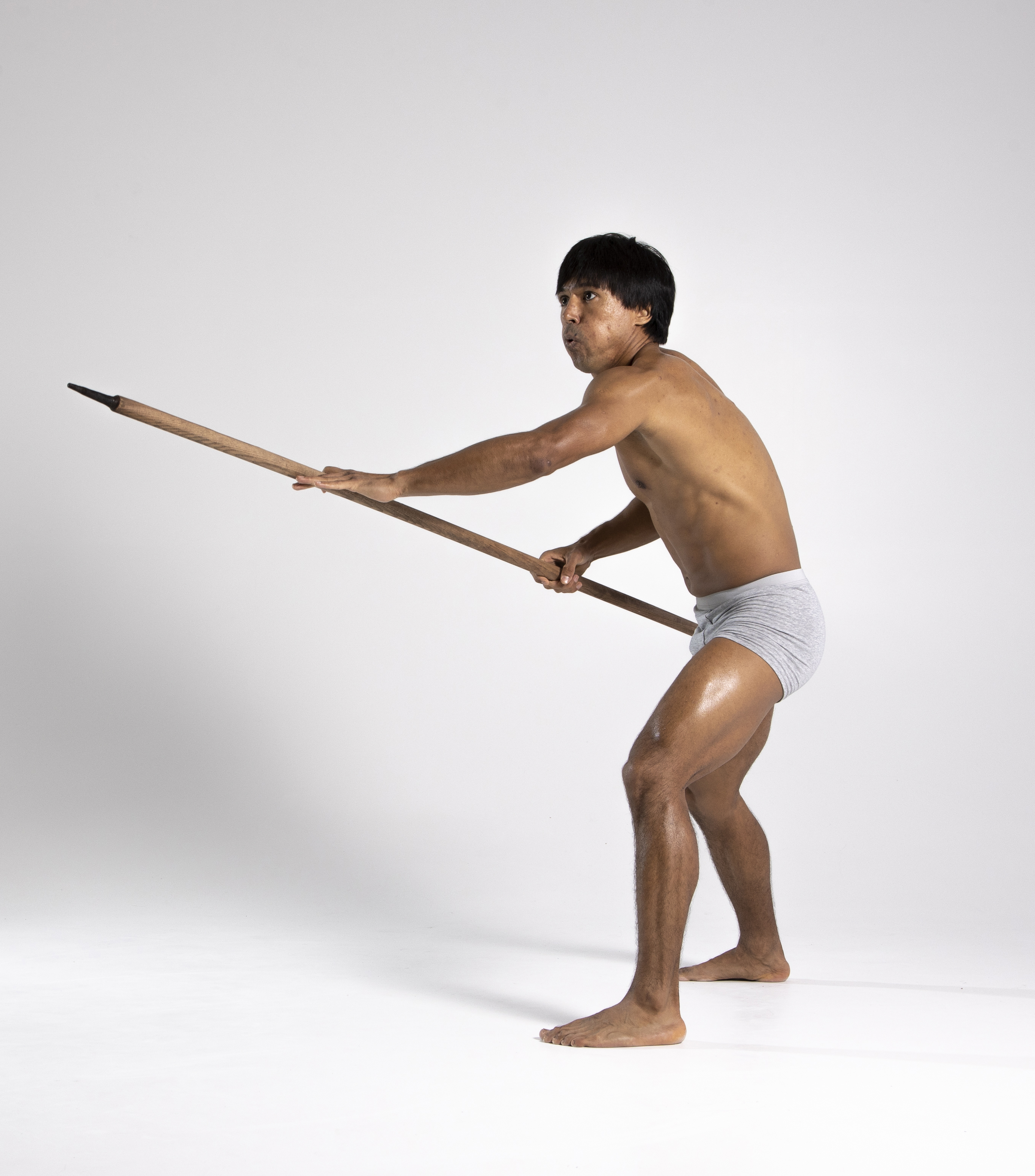 a barechested man in grey underwear holds a spear in a dynamic pose, set against a white background. he is positioned in a stance suggesting action or readiness, possibly conveying the image of a warrior or indigenous hunter.