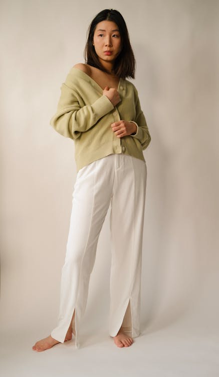 a full shot of an asian woman standing barefoot indoors, wearing a light green cardigan and white slit pants. she's positioned in front of a lightcolored backdrop, posing with a slightly offtheshoulder look.