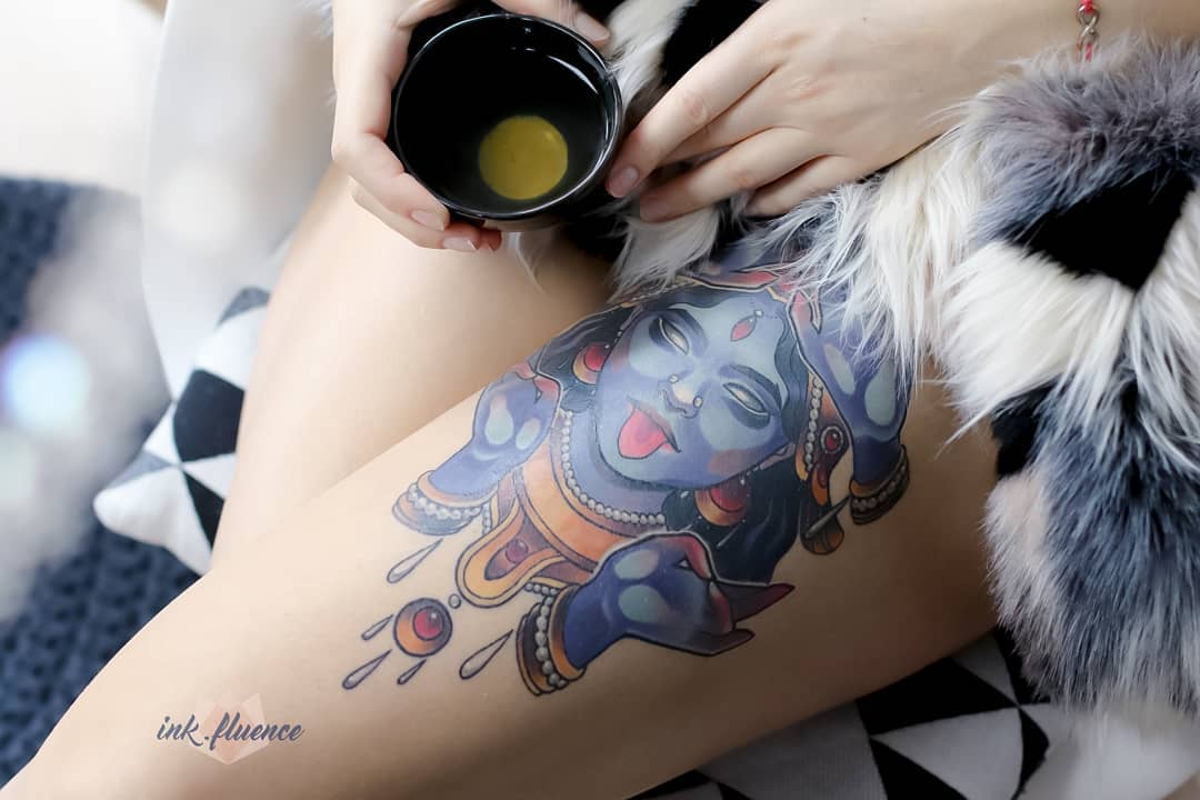 the image shows a closeup of a woman's upper thigh. she is wearing a faux fur coat and holding a cup of tea. the focus is on her colorful tattoo of a hindu deity, possibly kali, depicted in blue and gold hues. the tattoo is highly detailed and spans a significant portion of her thigh. the lighting is soft and natural, creating a warm and inviting atmosphere.