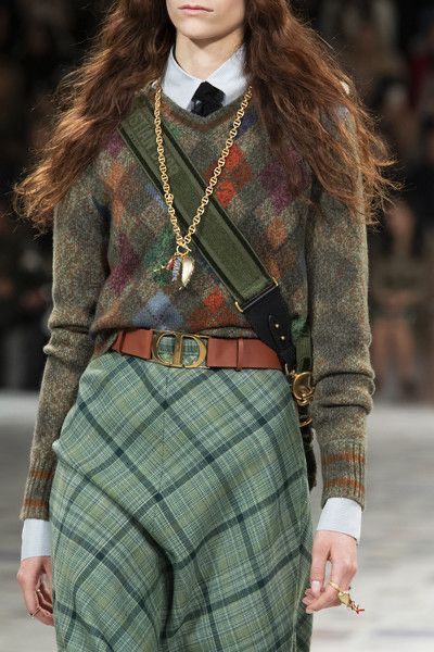 loseup view of a fashion model showcasing an autumnal outfit on a runway. the model is wearing a green and brown patterned sweater, a green and brown plaid skirt, a brown leather belt with a dior buckle, a gold necklace with a pendant, and a green and brown shoulder bag. the model's long brown hair is loose and flowing, completing the look. the overall style suggests a blend of classic and modern elements, with an emphasis on earthy tones and textures. the image is welllit and the focus is sharp, making the details of the outfit clearly visible.