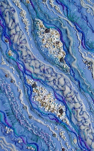 a closeup shot of an abstract blue and white needlework art piece with various embellishments. the main color is blue with white and light blue embellishments. the artwork is done in a variety of textures and patterns. there are beads, sparkles, and other embellishments throughout.  this is a handmade, decorative artwork piece.
