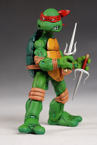 a closeup studio shot of a raphael teenage mutant ninja turtle action figure. he is green with a red headband, orange shell, and holding two silver sai weapons. the background is a neutral gray.