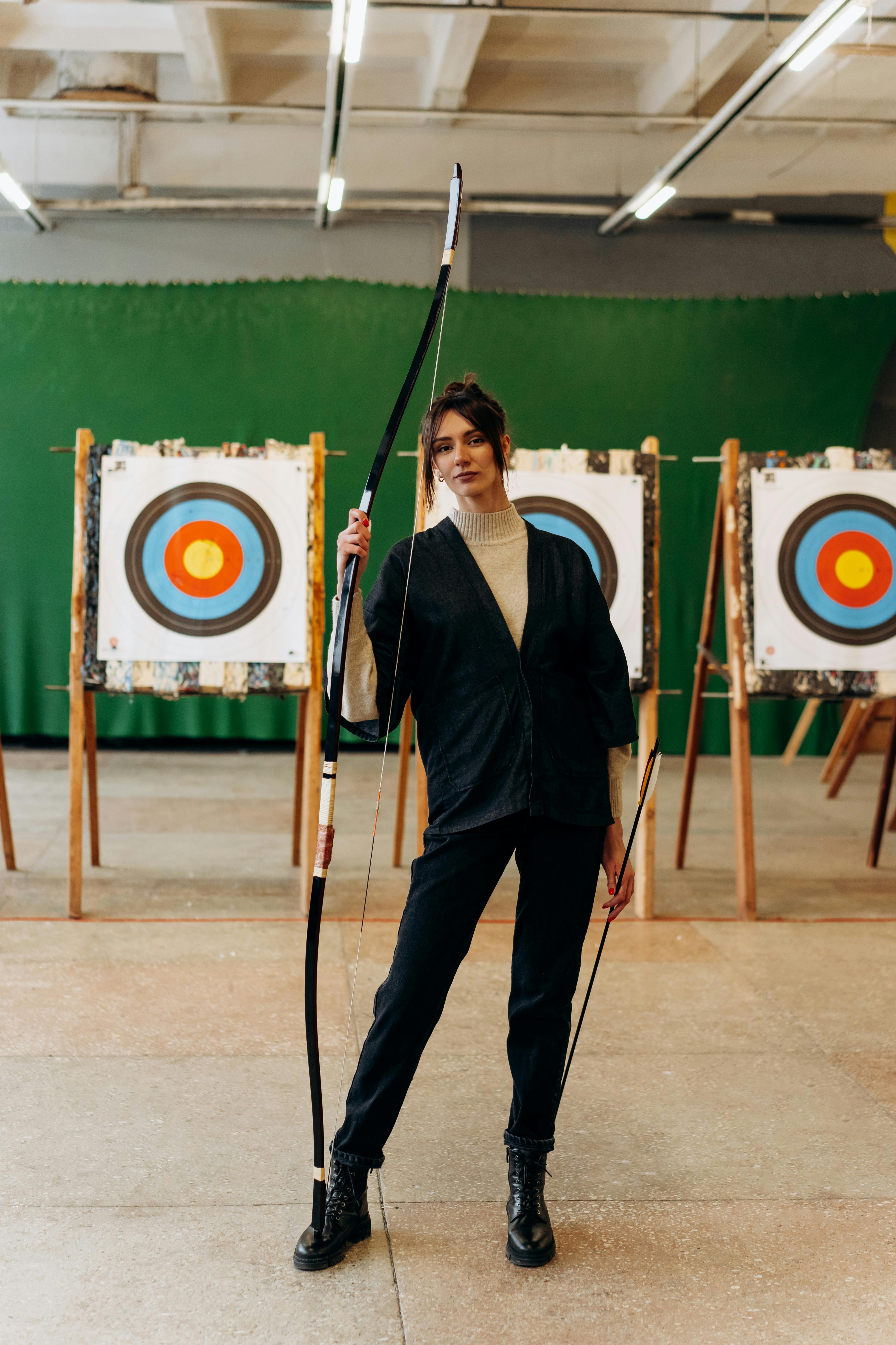 a young woman stands in an indoor archery range, holding a bow and arrow.  she is dressed in black pants and a black jacket over a cream colored sweater.  she is looking at the camera and has a serious expression on her face.  there are three target stands behind her with standard archery targets on them. the floor is light colored, and the walls are green. the overall atmosphere suggests a practice session or preparation for a competition.