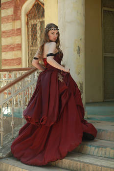 a fulllength shot of a woman in a long burgundy gown, standing on a staircase in front of an old building. the dress is styled in a victorian fashion with layers and a corset. the woman has a crown on her head and looks directly at the viewer.