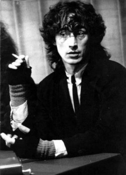 a black and white portrait of a man in a suit, smoking a cigarette. he is looking directly at the camera with a serious expression on his face. the photo is taken from a low angle, making the man appear larger than life. the man is wearing a suit jacket, a button up shirt, and a tie. he is also wearing a glove on his left hand. the man's hair is long and styled in a messy fashion. the background is blurred, which helps to focus attention on the man. the image evokes a feeling of coolness and intensity.