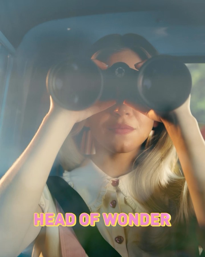 the image shows a blonde woman sitting in a car, holding binoculars up to her eyes.  she appears curious and perhaps engaged in some sort of observation or investigation. the scene is slightly blurry and sunlit, creating a soft and mysterious atmosphere. the text "head of wonder" overlays the image in a stylized pastel font.  the overall aesthetic suggests a film still or movie poster, possibly for a drama or mystery.