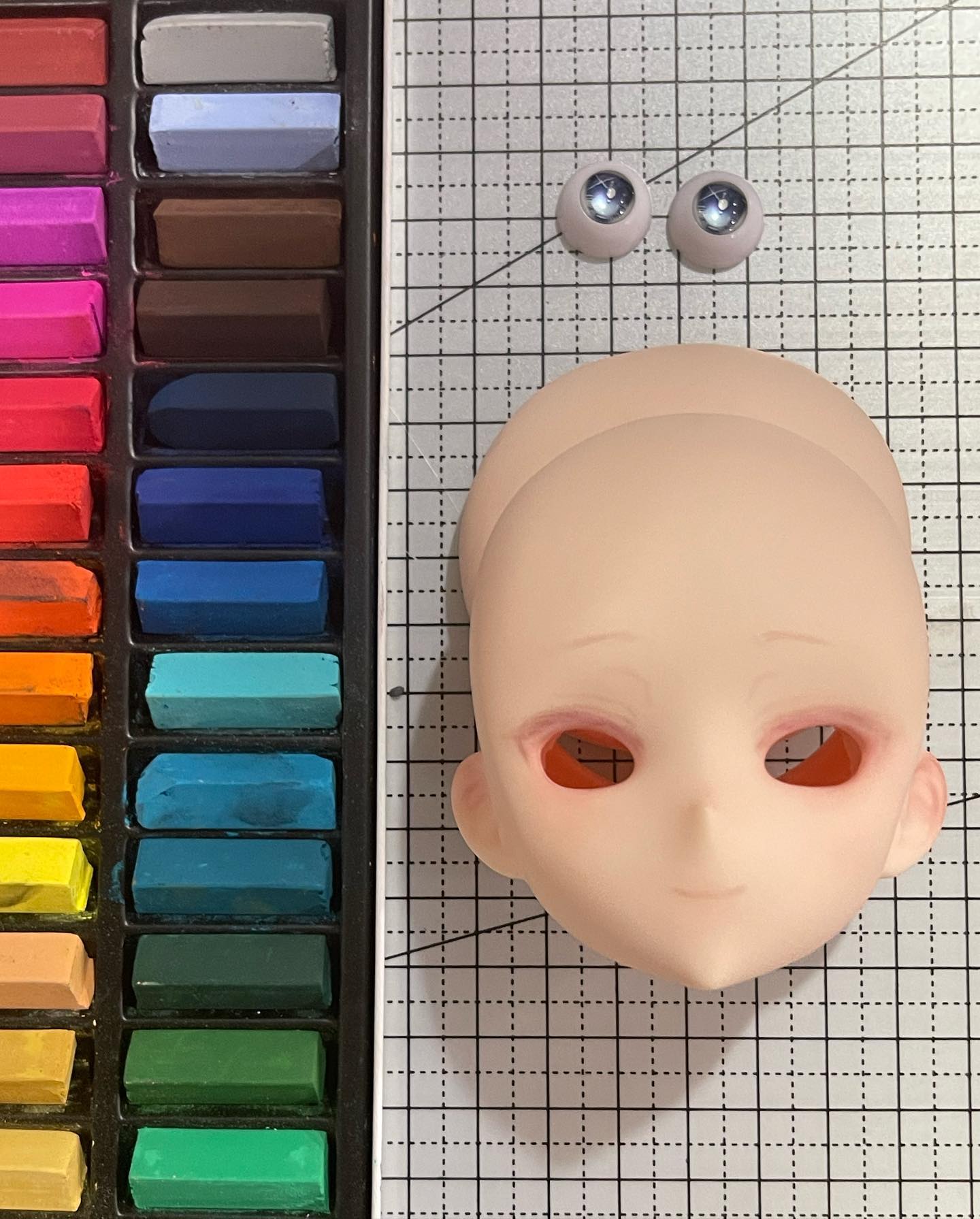 a doll head lays on a gridded craft mat next to a set of pastels. the doll head is painted with blushing cheeks and pink lips. a set of silver doll eyes lay above the doll head.  the image is lit from above.