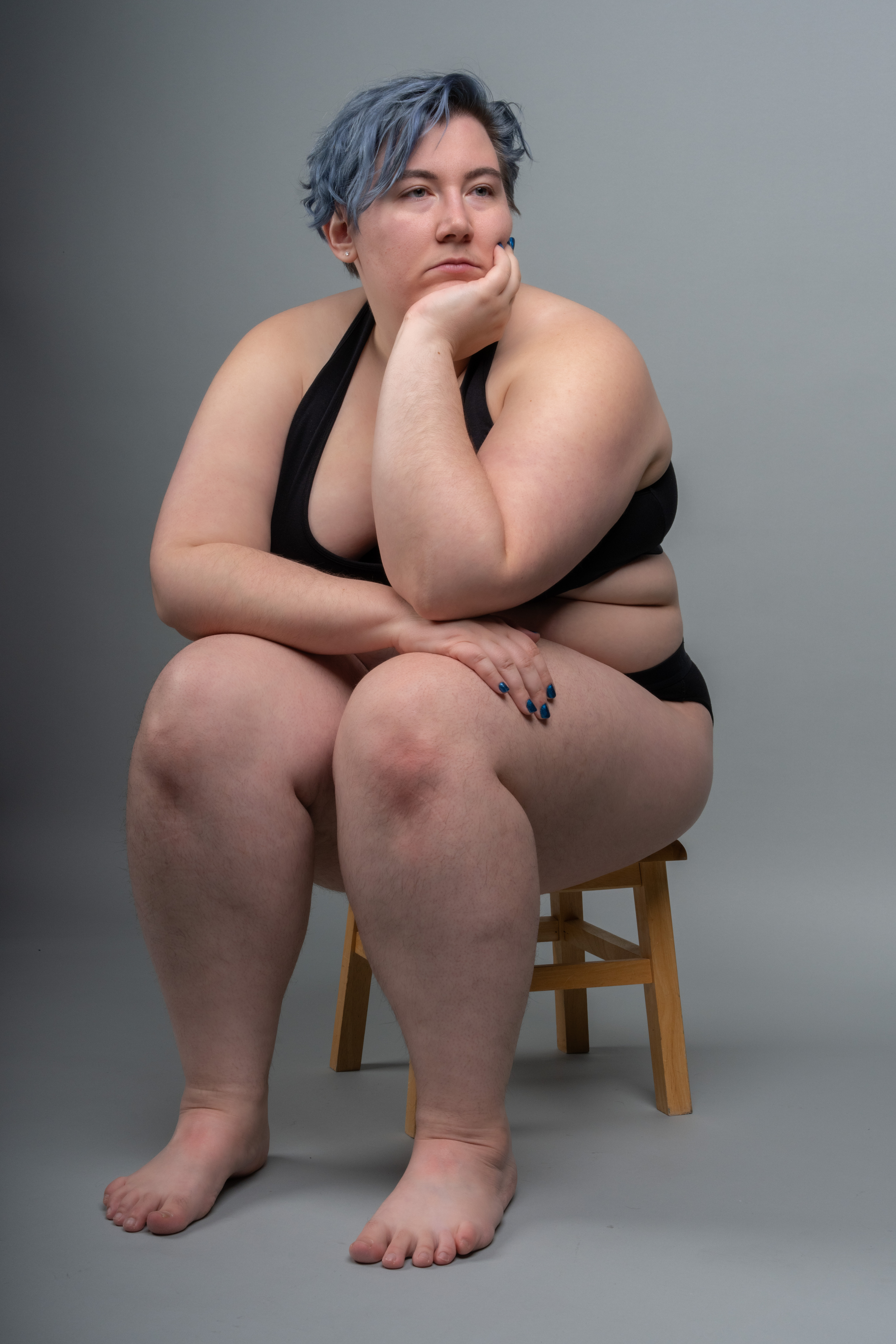 a plussize woman with blue hair is sitting on a wooden stool in a studio setting. she is wearing a black swimsuit and has a thoughtful expression on her face, resting her head on her hand. the background is a plain gray. she is barefoot.