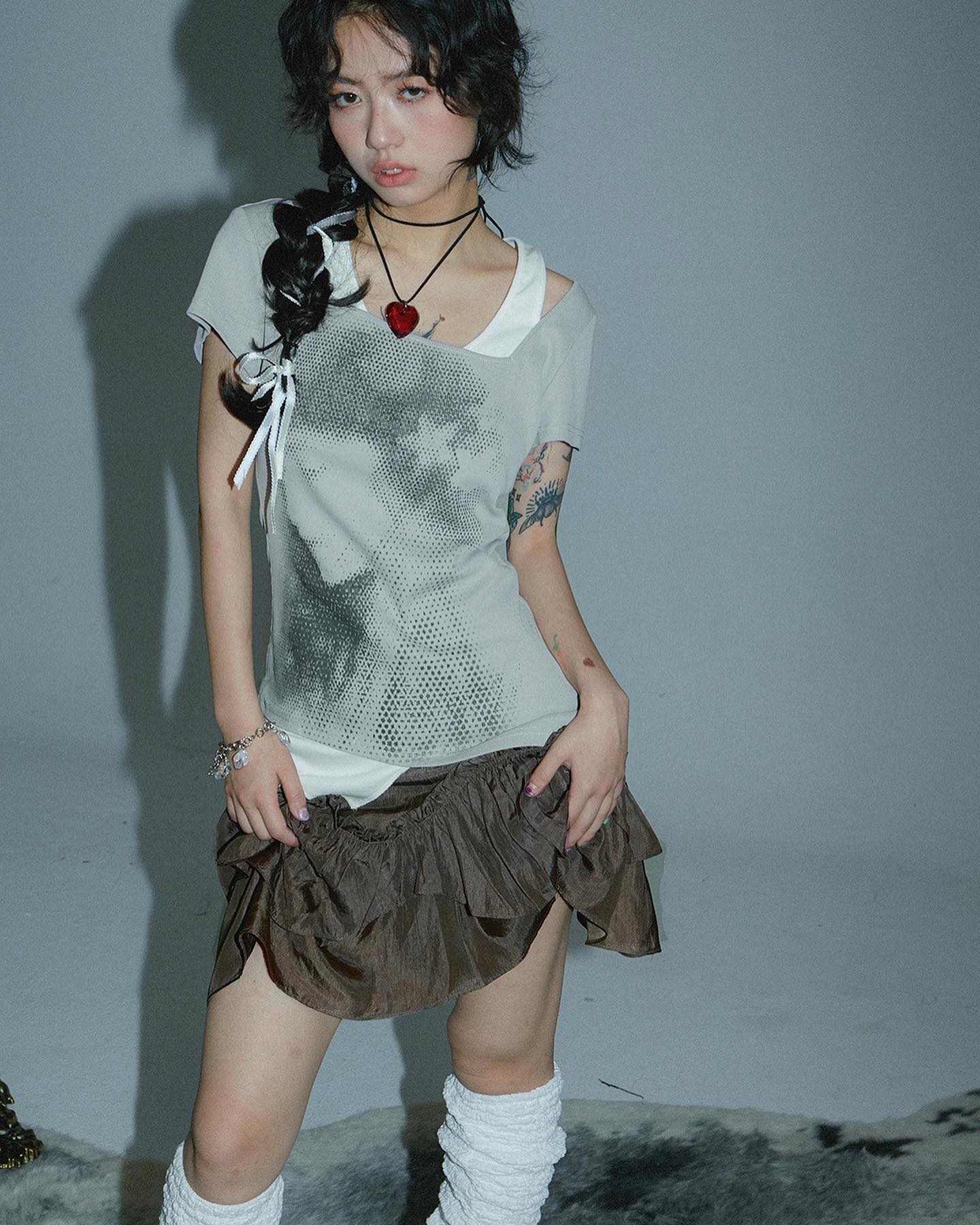 a young asian woman with dark hair styled in braids is posing in a studio against a grey background. she is wearing a grey graphic tshirt with a distressed neckline, a brown ruffled mini skirt, white leg warmers, and several accessories including a heartshaped necklace.  she has subtle makeup and small tattoos visible on her arm.  the overall style is trendy and slightly edgy.