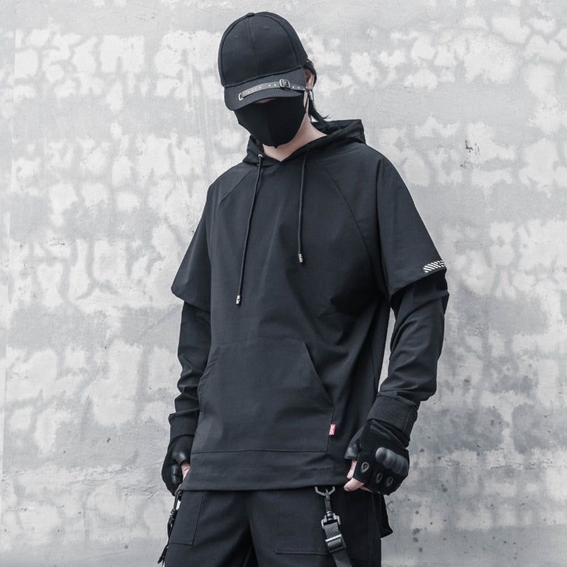 a man is wearing a black hoodie with a black cap and gloves. he is posing in front of a gray wall. the man is wearing a streetwear look.
