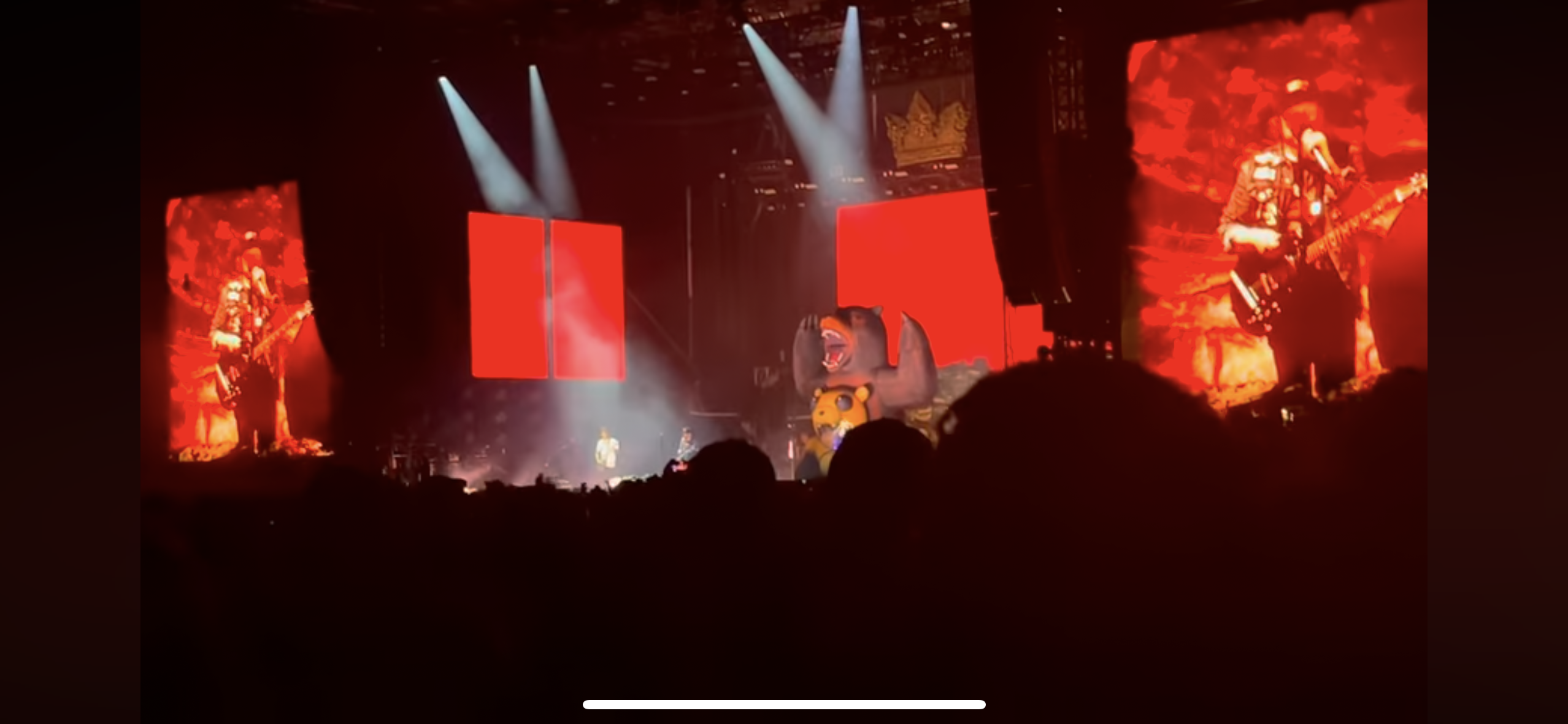 a concert scene with a large crowd of people gathered in front of a stage lit by spotlights. the stage features two large screens displaying images of musicians, one of them playing a guitar. there is a bear inflatable in the center of the stage. the crowd is facing the stage with their arms in the air, cheering and singing along to the music.