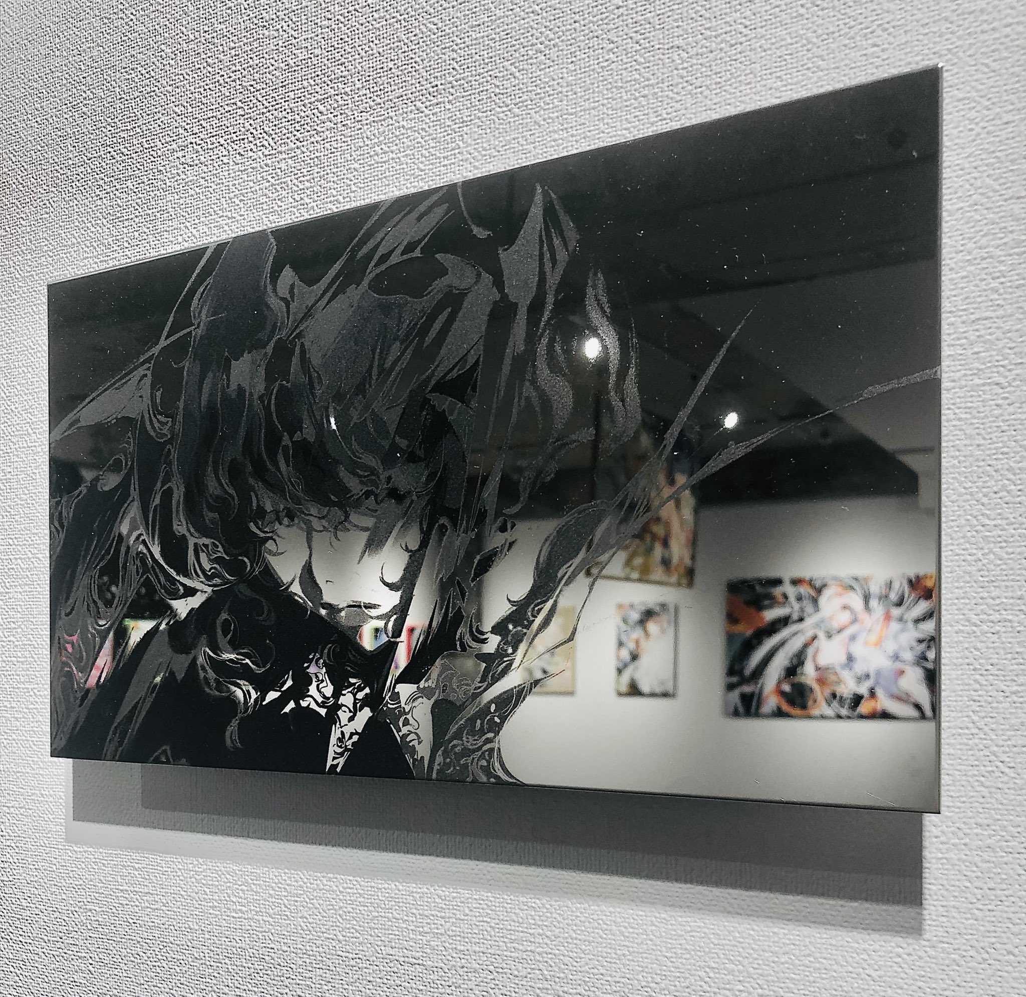 the image showcases a sleek, black rectangular artwork mounted on a wall. it appears to be a mirror with an intricate illustration etched onto its surface. the illustration, rendered in black and white, depicts a stylized character, possibly from anime or manga, with flowing hair and sharp, dynamic lines. the mirror reflects the opposite wall of the gallery, offering glimpses of other framed artworks, suggesting this piece is part of an art exhibition. the lighting is soft and diffused, highlighting the glossy surface of the mirror and the details of the etched illustration.