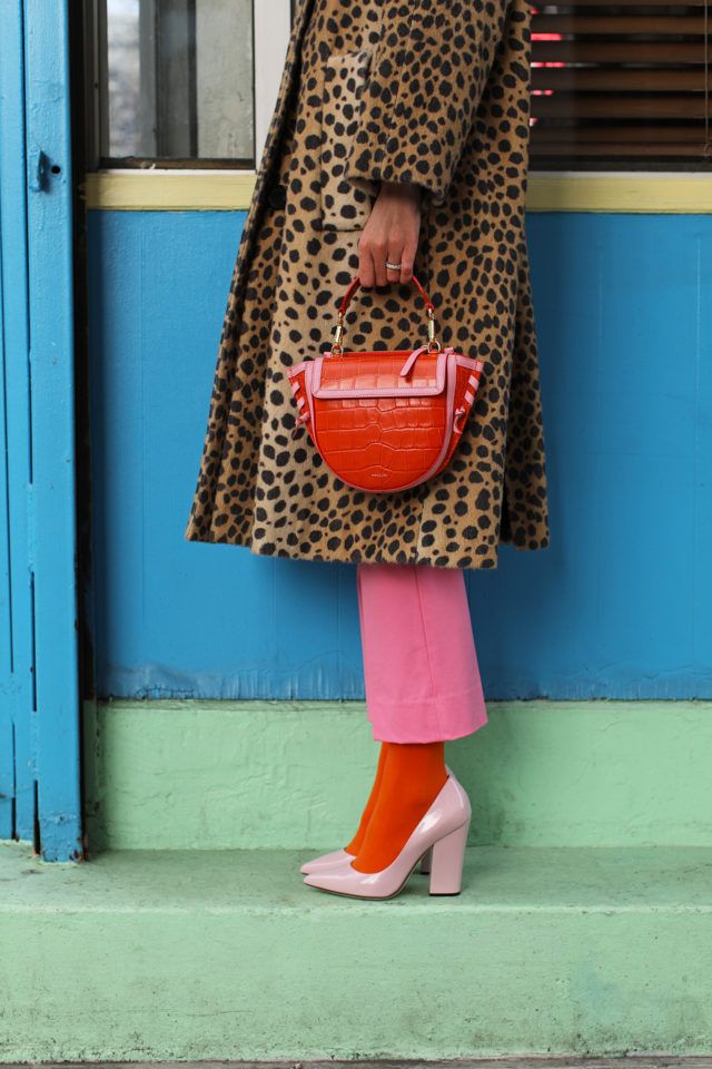 a woman is standing against a blue and green wall wearing a leopard print coat, pink pants, orange socks, and pink heels. she is carrying an orange handbag. the overall style is colorful and fashionable.