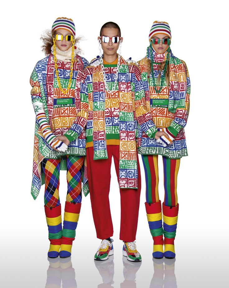 the image shows a group photo of three models wearing vibrant, multicolored outfits. they are dressed in outerwear with a square pattern, hats, sunglasses, scarves, and coordinating bottoms, all in rainbow colors. the attire includes knitwear and winter clothing, representing a quirky and unique fashion style, likely from the united colors of benetton. the models are posing in a studio setting.