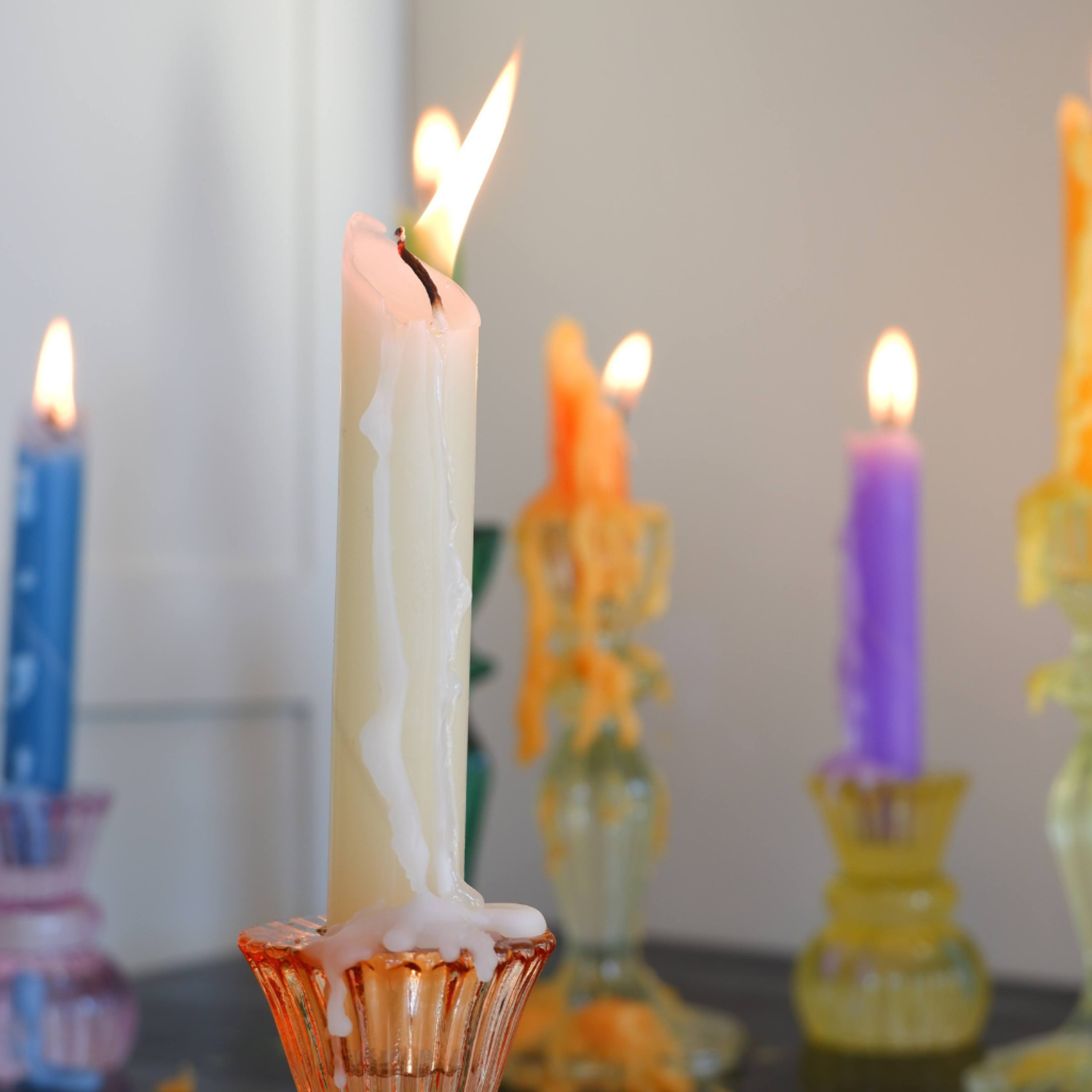 a close up of five lit candles, the center candle is white, two are orange, one is blue and one is purple. the candles are in various candle holders and sitting on a table, the image is blurred in the background.