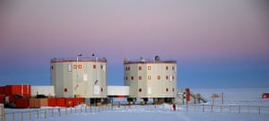 a wideshot landscape photo showing two industrial style buildings in a snowy, icy arctic or antarctic setting. the buildings are rectangular, metal, with windows and doors. the buildings are in a remote, wilderness area, perhaps a science research station. there is a fence in front of the buildings, and the horizon has a blue sky with clouds. the ground is covered in white snow and ice, making the landscape look very cold.